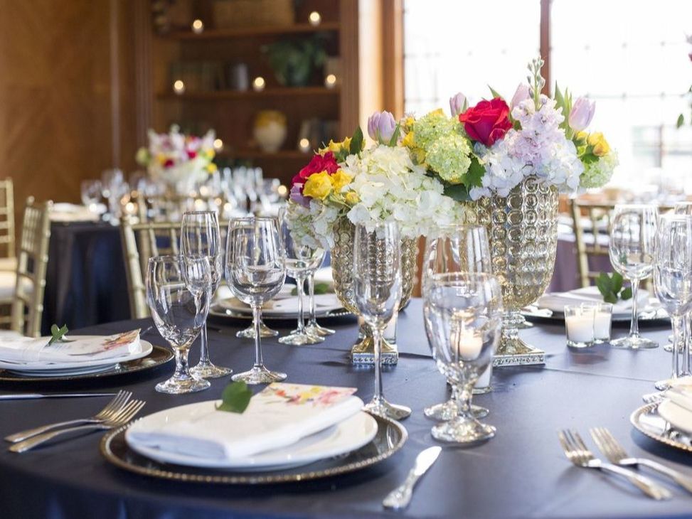 A well appointed place setting with full glass glasses and a large colorful flower centerpiece